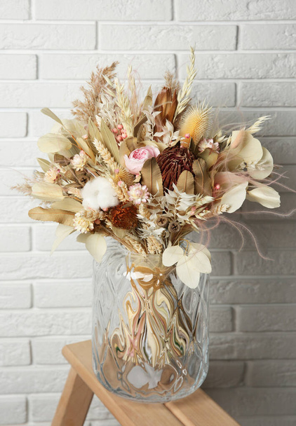 Beautiful dried flower bouquet in glass vase on wooden ladder near white brick wall - Fotó, kép