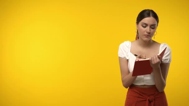 Pensive woman writing on notebook isolated on yellow - Footage, Video