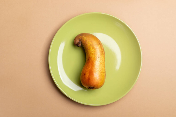 View from above to one ugly crooked ripe pear lying on round green plate on beige background. - Foto, Imagem