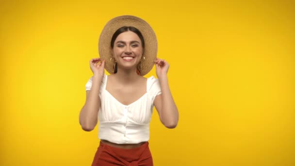 Usmívající se žena ve slunečním klobouku při pohledu na kameru izolované na žluté - Záběry, video