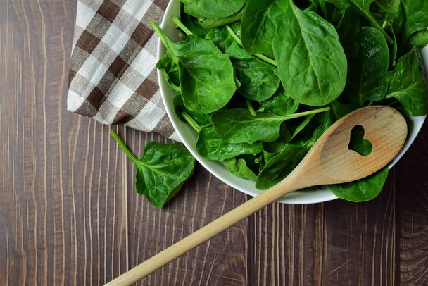 Healthy food. Spinach leaves, in a white bowl and on a checkered cloth, wooden spoon. Zenith view. Copy space. - Foto, imagen