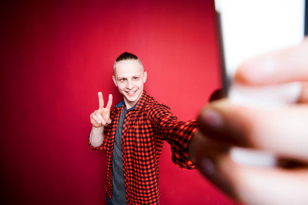Atractivo hombre sosteniendo un teléfono en su mano y tomando fotos de sí mismo mostrando señal de paz con sus dedos - Foto, imagen