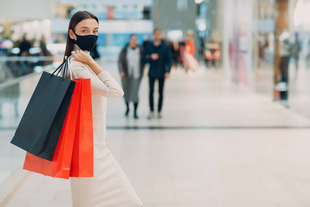 Молодая взрослая женщина в защитной маске для лица несет бумажные пакеты в руках в торговом центре - Фото, изображение