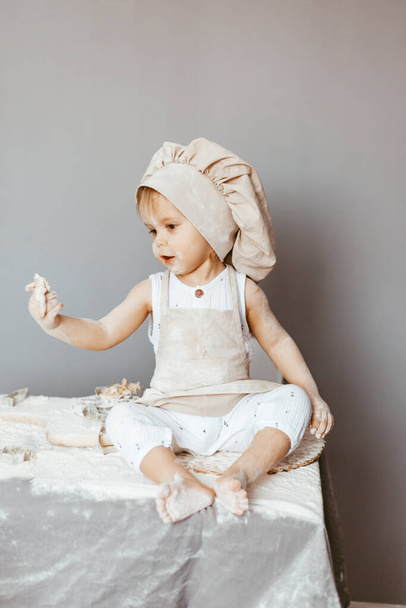 a happy child in an apron and a chef's hat prepares dough, bakes cookies in the kitchen - Фото, изображение