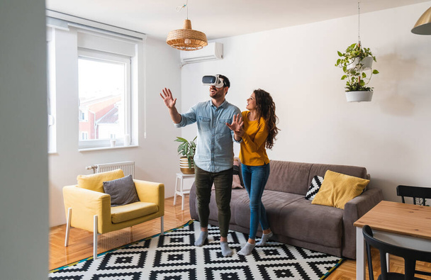 Happy couple using VR headset at home. Amazed man touching the air during the VR experience and woman holds him - Photo, Image