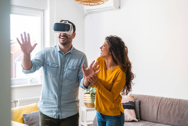 Young couple enjoying having fun with virtual reality headset. Man touching the air during the VR experience and woman holds him - Foto, Imagen