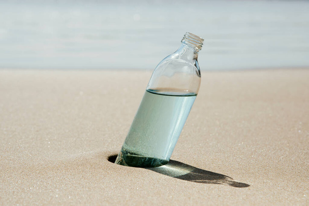 a glass reusable water bottle on the wet sand of the seashore of a lonely beach, with the sea in the background - Foto, imagen