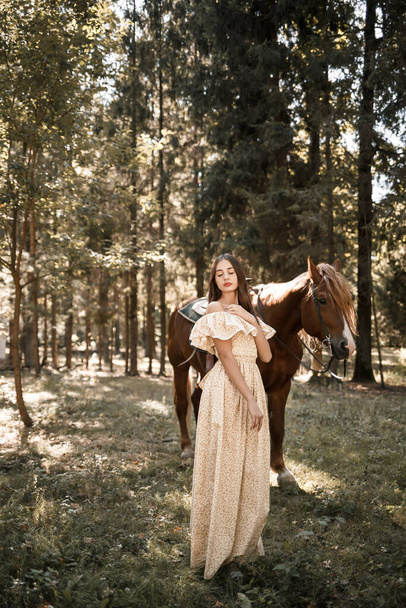 A beautiful young girl dressed in a dress stands near a horse in the forest - Фото, изображение