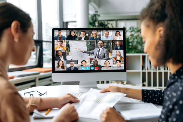 Online-Geschäftstreffen, Brainstorming. Blick über die Schultern zweier Frauen auf einen Computerbildschirm mit verschiedenen erfolgreichen multirassischen Menschen, die sich in einer Videokonferenz versammelt haben, um Arbeitsfragen zu diskutieren - Foto, Bild