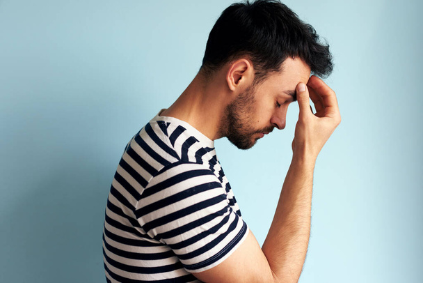 Side view of a tired overworked young man keeps hand on forehead with closed eyes, dressed in striped t-shirt, isolated over blue studio wall. A male has a headache. - Фото, изображение