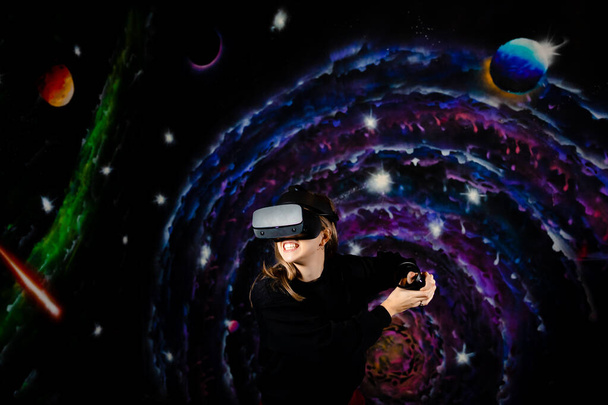 A young girl plays battles in a video game wearing VR virtual reality glasses. Space background, stars and planets.  - Foto, afbeelding