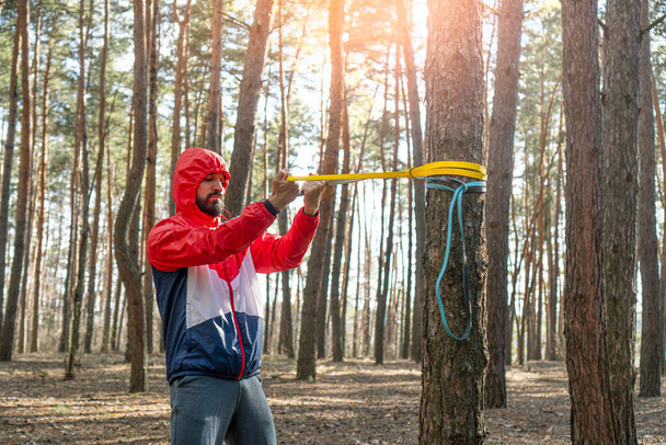 Athletic bearded man pulls the resistance band in a morning forest.  - 写真・画像