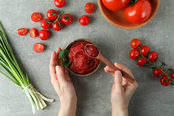 Vrouwelijke handen houden kom en lepel met tomatenpasta op grijze achtergrond met ingrediënten - Foto, afbeelding