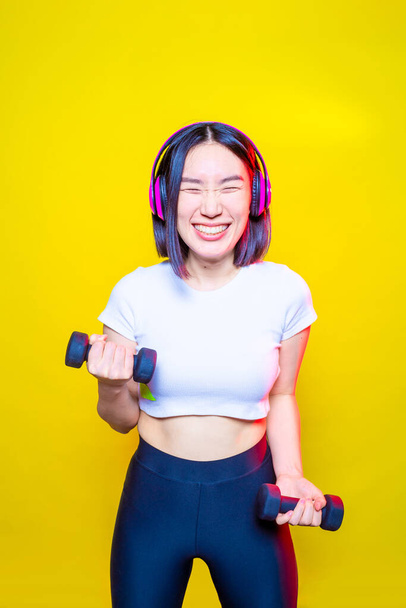 Adatto giovane donna asiatica sollevamento pesi su sfondo giallo ascoltare musica divertendosi - Foto, immagini