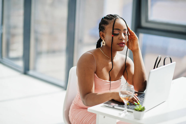 Fashionable photo of young beautiful romantic african american woman with glass of wine, wearing pink dress working with laptop. - Zdjęcie, obraz