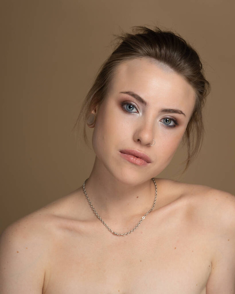 Open-eyed girl with heterochromia, nose piercing and plug in one ear, and strange hairstyle. With amazing professional makeup and silver chain around her neck. Beige background. Studio shot - Photo, Image