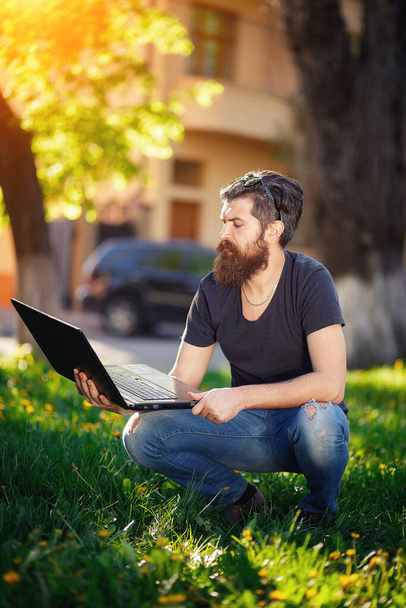 Brodaty programista z długą brodą i ciemnymi siwymi włosami w koszuli i dżinsach przykucnął na zielonej trawie w parku z laptopem. Twórca gry spoczywa w naturze - Zdjęcie, obraz