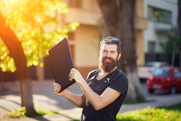 Cheerful bearded male programmer with a long beard and dark gray hair in a shirt and jeans waving a laptop against the background of a green park in spring - Photo, Image