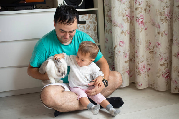 adorable baby sits in dad's arms and strokes a decorative rabbit. father shows little child easter bunny. domestic animals in a family with children - Photo, Image