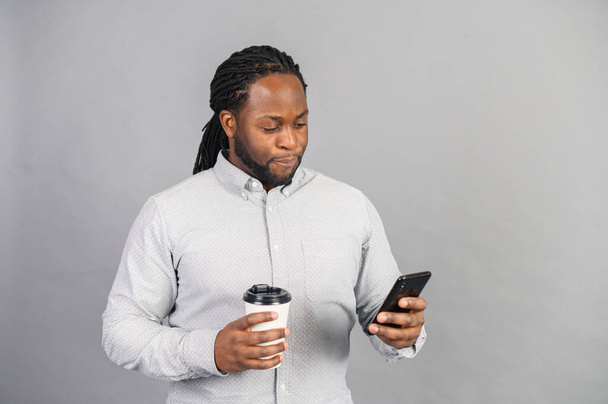 Smiling African-American man holding phone and cofee, isolated on grey - Photo, image