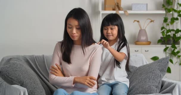 Offended asian woman pretends to be angry because of her daughters bad behavior ignores little girl sits with her back to child worries about quarrel cute caring kid strokes mom on shoulder consoles - Footage, Video