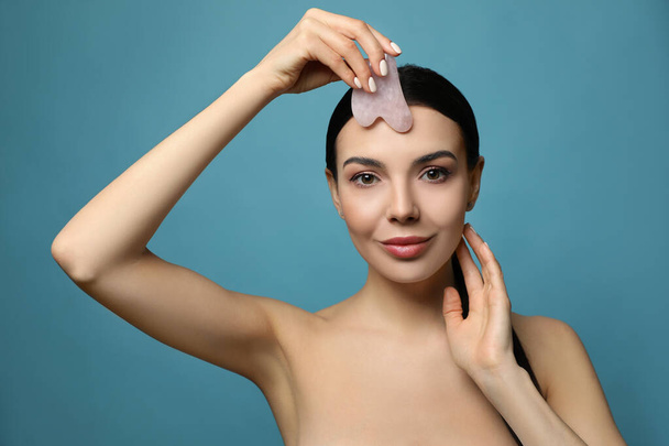 Beautiful young woman doing facial massage with gua sha tool on blue background - Photo, Image