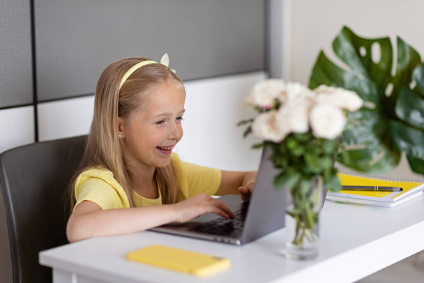 Cute little caucasian girl with blonde hair in fashionable dress illuminating yellow color sitting at home during coronavirus pandemic quarantine and using laptop and mobile phone. Stay at home during - Фото, изображение