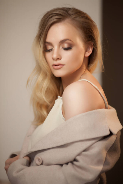 Portrait of a blonde woman wearing beige coat, looking over shoulder. Beautiful woman with everyday makeup. - Foto, imagen