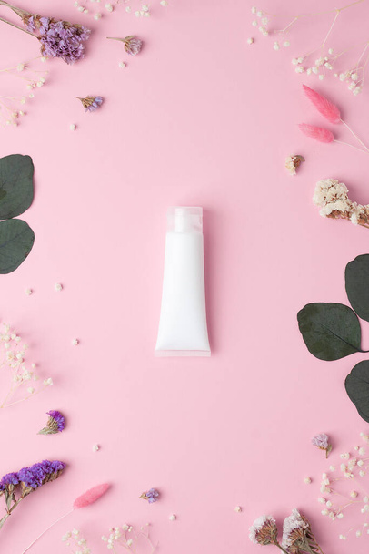 White tube of cosmetic cream with flowers and green leaves on pink background. Flat lay - Photo, Image