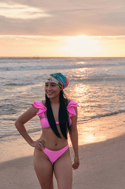 Латиноамериканка в розовом летнем купальнике и бандане на берегу моря с закатом на спине - Фото, изображение