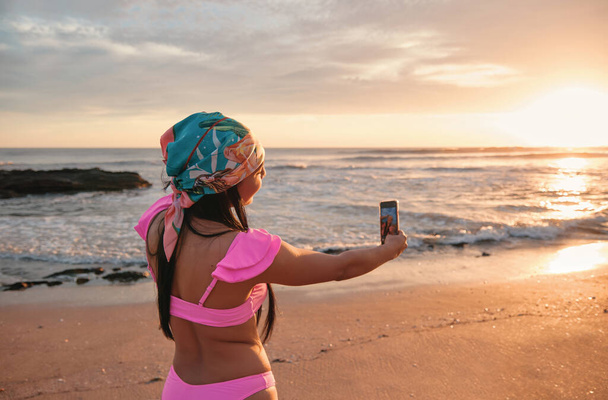 Роскошная латиноамериканка в розовом бикини и бандане, делающая селфи со смартфоном в красивом закате на берегу моря. - Фото, изображение