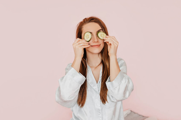 Girl make homemade face beauty masks. Cucumbers for the freshness of the skin around the eyes. Woman take care of youthful skin. Model laughing and having fun in spa on pink background - Fotoğraf, Görsel