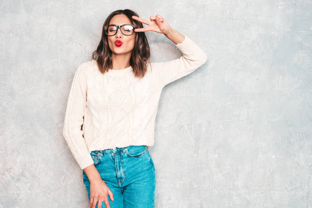 Young beautiful smiling woman. Trendy female in casual summer jeans clothes. Positive female with red lips shows facial emotions. Funny and cheerful model posing near gray wall in studio in eyewear - Photo, Image