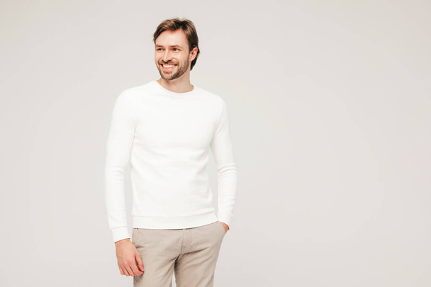 Portrait of handsome smiling hipster lumbersexual businessman model wearing casual white sweater and trousers. Fashion stylish man posing against grey background in studio - 写真・画像
