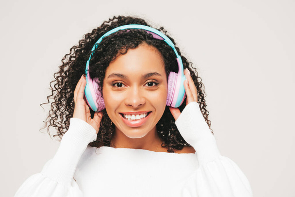 Beautiful black woman with afro curls hairstyle.Smiling model in sweater and jeans.Sexy carefree female listening music in wireless headphones.Posing in studio on white background - Fotó, kép
