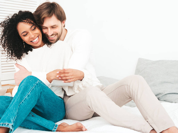 Smiling beautiful woman and her handsome boyfriend. Happy cheerful multiracial family having tender moments. Multiethnic models lying in bed and hugging in white interior. Embracing each other - 写真・画像