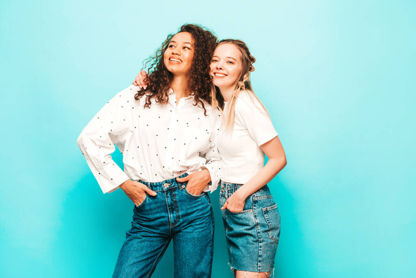 Dos jóvenes hermosas y sonrientes hipsters internacionales con ropa de verano de moda. Sexy mujeres despreocupadas posando cerca de la pared azul en el estudio. Modelos positivos divirtiéndose. Concepto de amistad - Foto, imagen