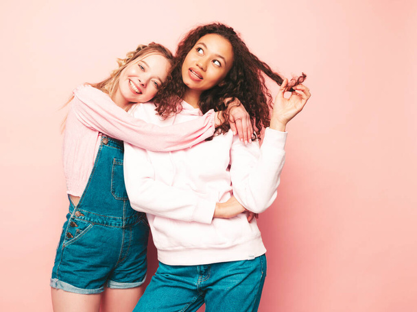 Dos jóvenes hermosas y sonrientes hipsters internacionales con ropa de verano de moda. Sexy mujeres despreocupadas posando cerca de la pared rosa en el estudio. Modelos positivos divirtiéndose. Concepto de amistad  - Foto, imagen