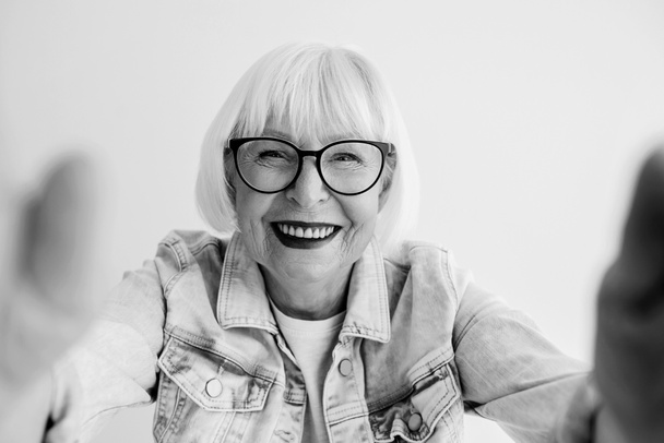 monochrome portrait of senior stylish woman with gray hair and in eye wear and denim jacket hugging you or making selfie. Love, hugs, anti age, positive vibes concept - Photo, Image