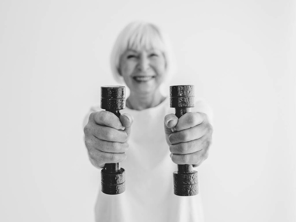 monochromatický portrét starší veselé ženy, která sportuje s činkami. Anti věk, sport, zdravý životní styl koncepce  - Fotografie, Obrázek
