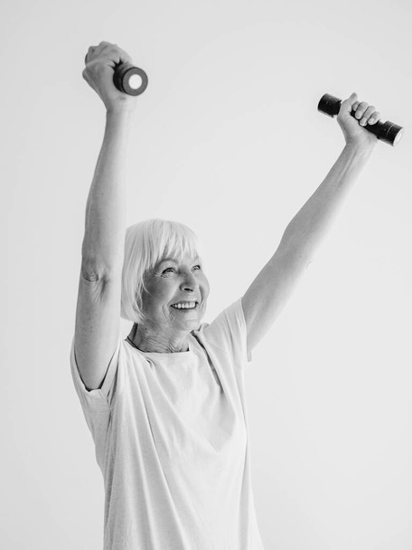 ダンベルとスポーツをしている先輩陽気な女性のモノクロの肖像画。抗年齢、スポーツ、健康的なライフスタイルの概念  - 写真・画像