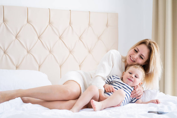 schöne Frau umarmt ihren niedlichen kleinen Sohn und lächelt, während sie auf dem Bett liegt. - Foto, Bild