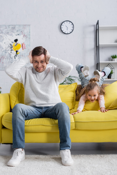 Smiling man covering ears near daughter on couch  - Foto, afbeelding