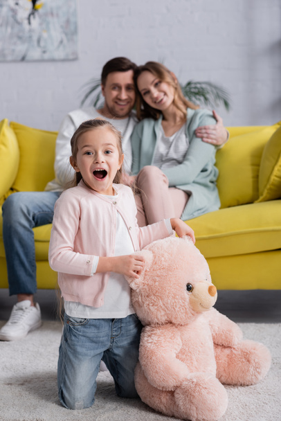 Niño sorprendido sosteniendo oso de peluche cerca de los padres en el sofá sobre fondo borroso  - Foto, imagen