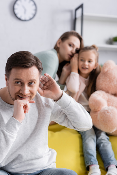 Un homme souriant écoute près de sa femme et de sa fille avec un jouet doux sur fond flou  - Photo, image