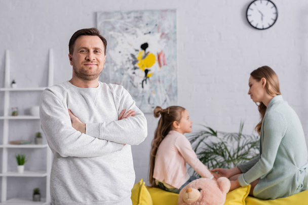 Hombre con brazos cruzados mirando a la cámara cerca de la familia borrosa en la sala de estar  - Foto, Imagen