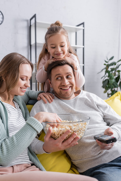 Smiling man with popcorn and remote controller sitting near wife and child  - Foto, Imagen