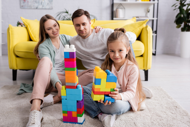 Kid with building blocks looking at camera while parents embracing at home  - Foto, imagen