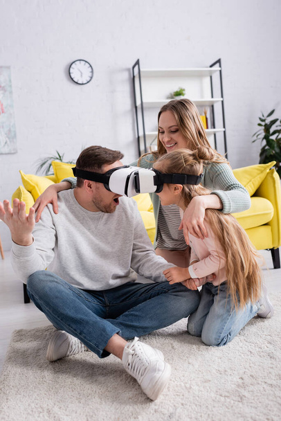 Father and daughter gaming in vr headsets near positive mother on carpet  - Foto, imagen