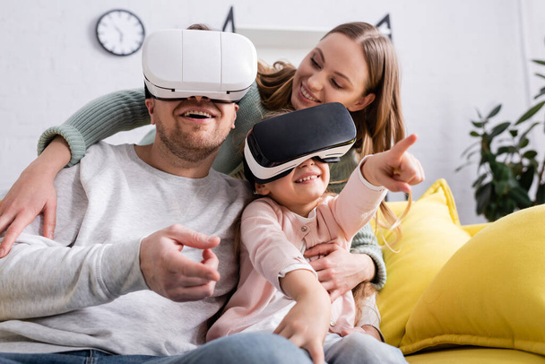 Smiling girl pointing with finger while playing in vr headset near parents  - Valokuva, kuva